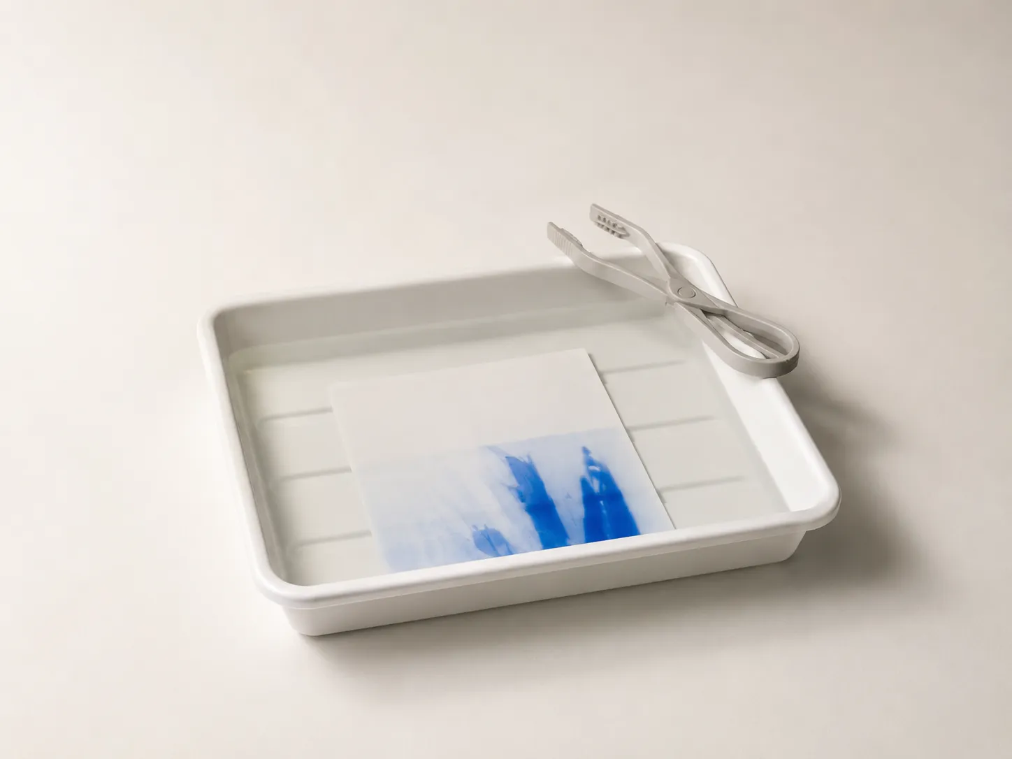 A print developing in a white darkroom tray, its blue areas only half visible, with print tongs resting on the rim