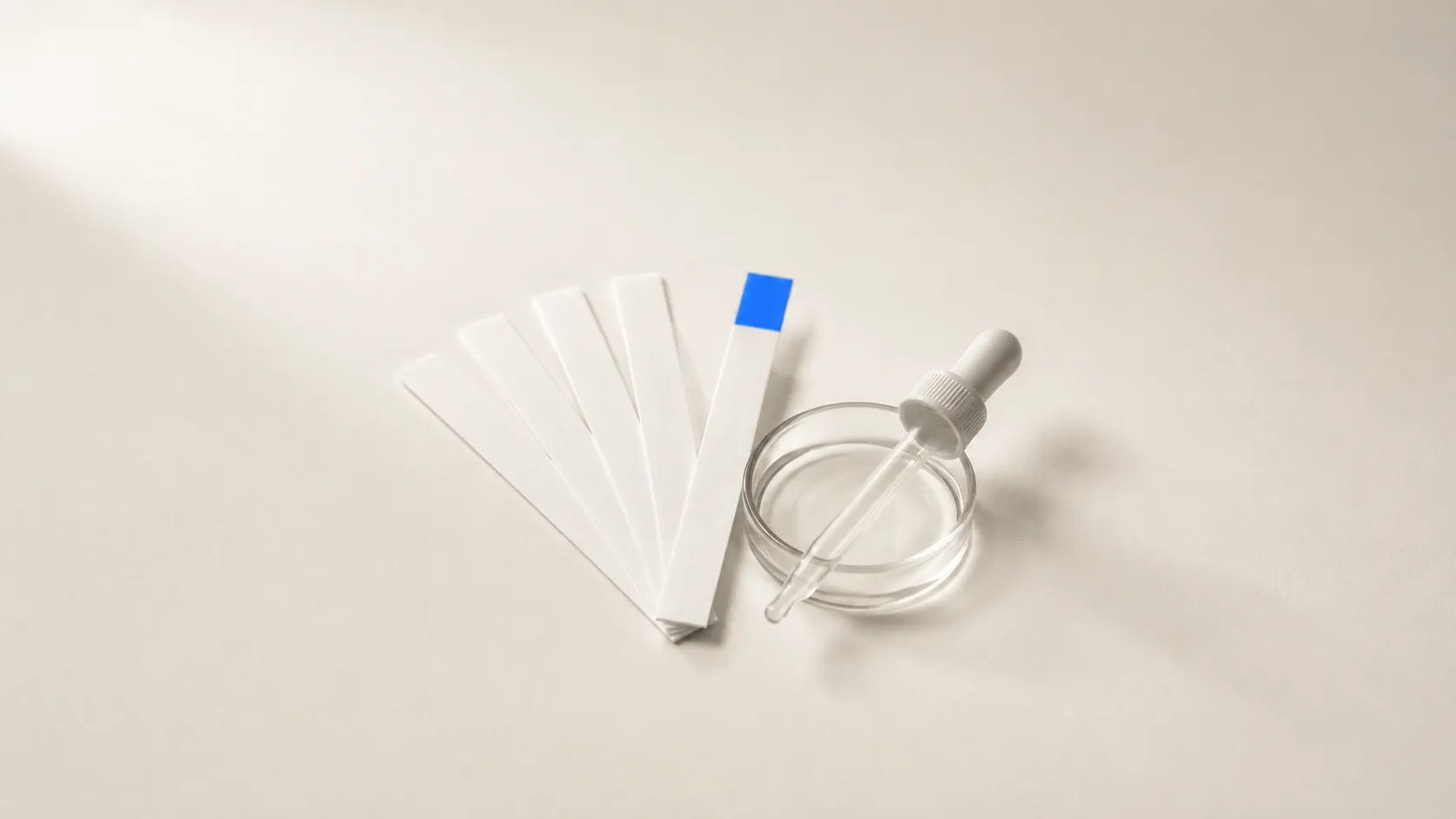 Five white test strips fanned out on a beige surface, the front one with a blue-stained field, next to a shallow glass dish with a pipette