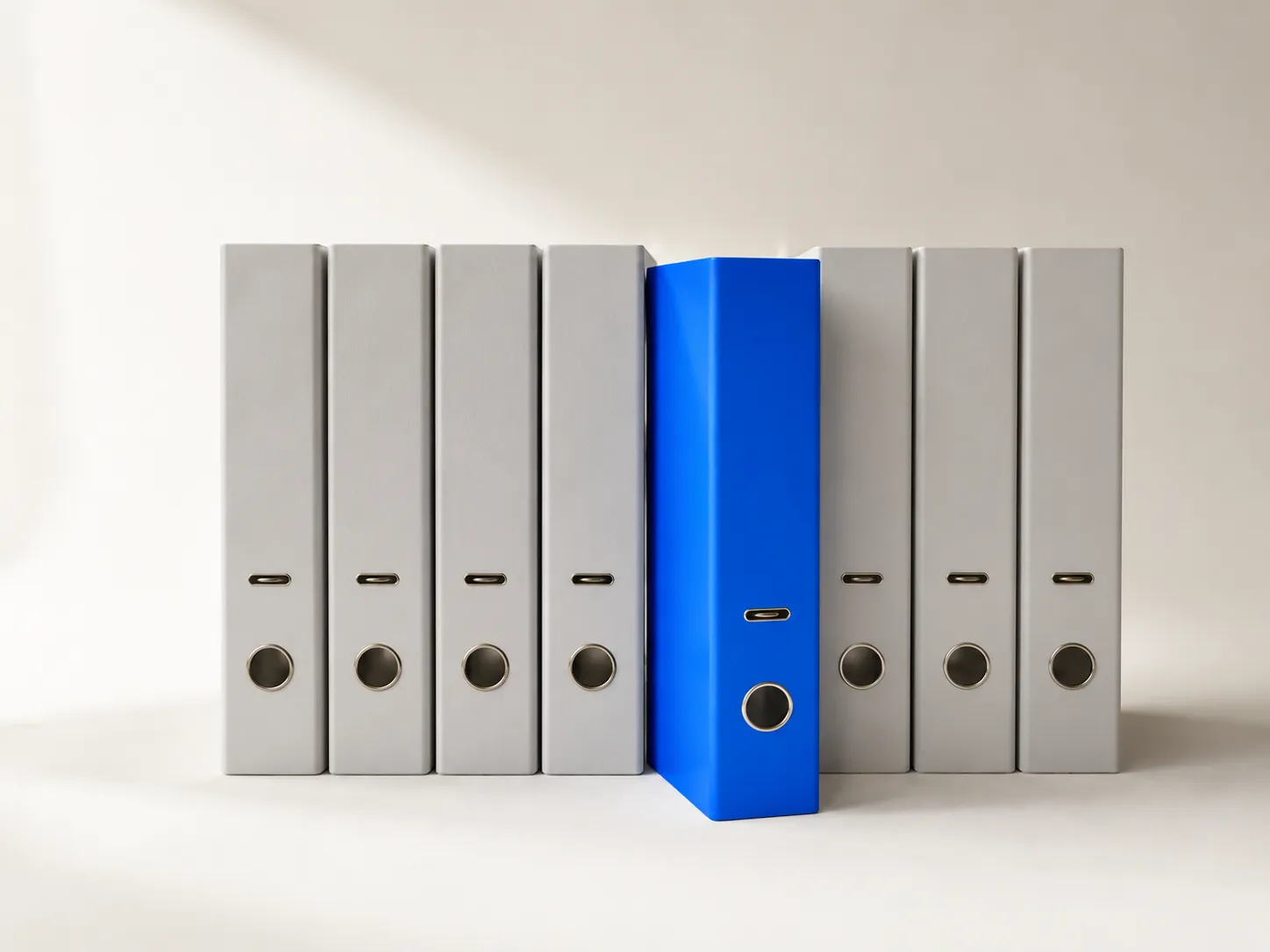 Eight ring binders side by side against a light background, seven grey and one blue standing slightly forward